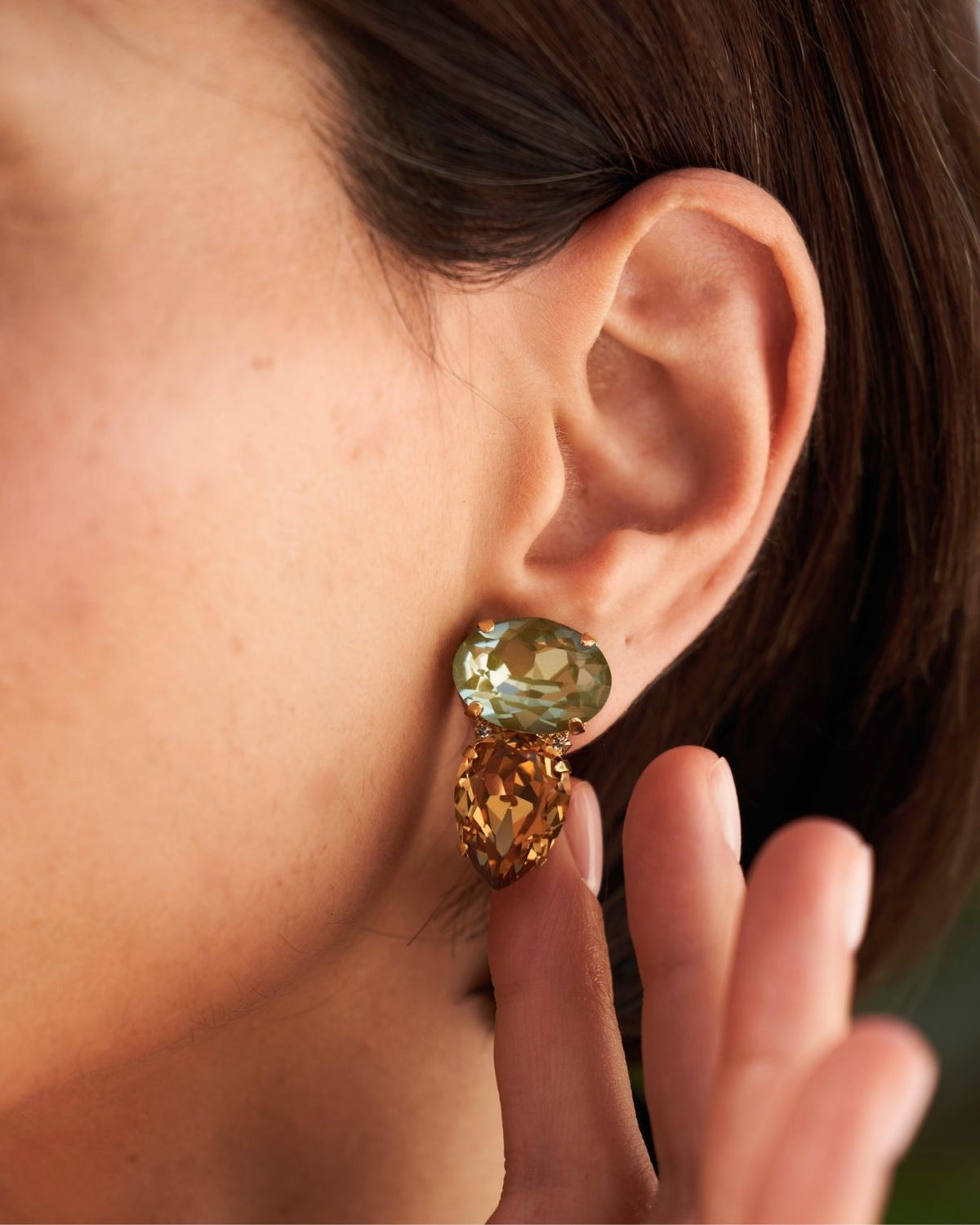 A close-up of a model's ear wearing a gold drop earring with a faceted light green top stone and a larger faceted amber-brown bottom stone.