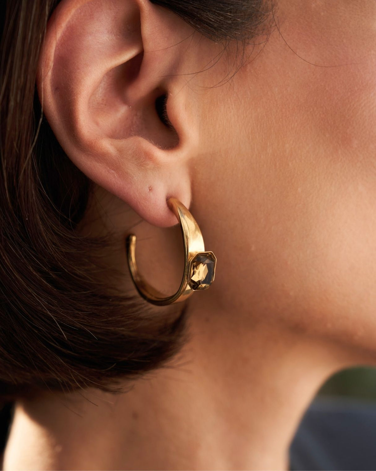 A close-up of a person's ear wearing a gold huggie hoop earring with a small square dark stone accent.