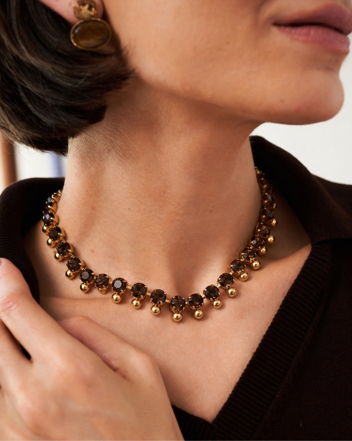 A close-up lifestyle shot of a model wearing a gold-plated collar necklace with dark brown smoky crystal accents, paired with a dark brown knit top.