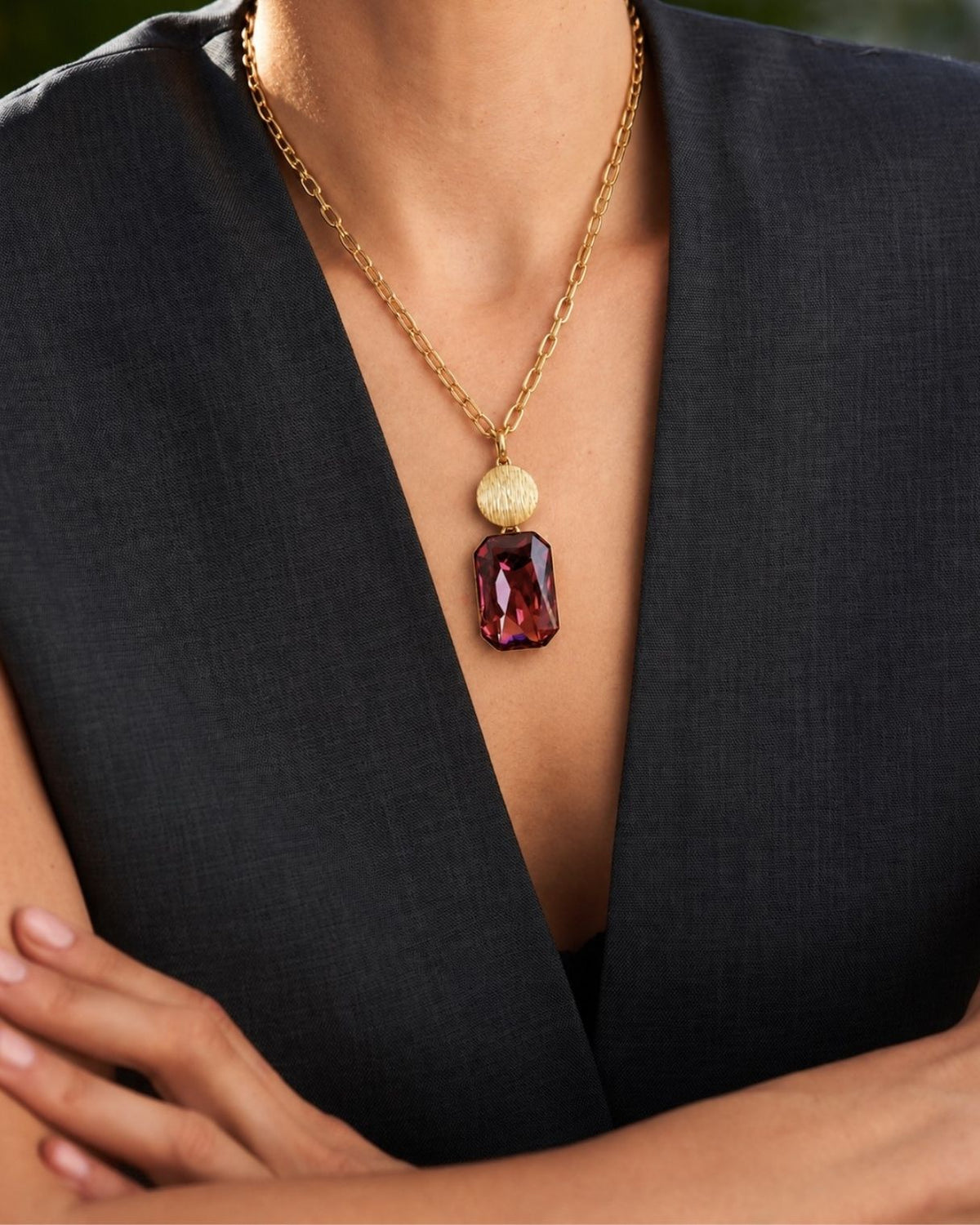A close-up of a model wearing a gold chain necklace with a textured gold disc and a large faceted burgundy gemstone pendant, styled with a dark gray sleeveless top.
