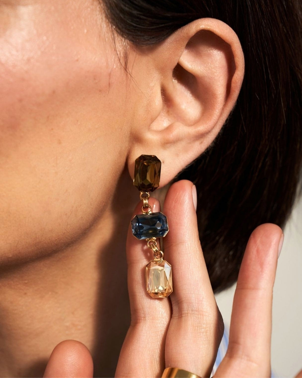 A close-up of a person's ear wearing a gold-tone drop earring with faceted dark brown, blue, and clear crystals, while their hand holds the earring in front of their face.