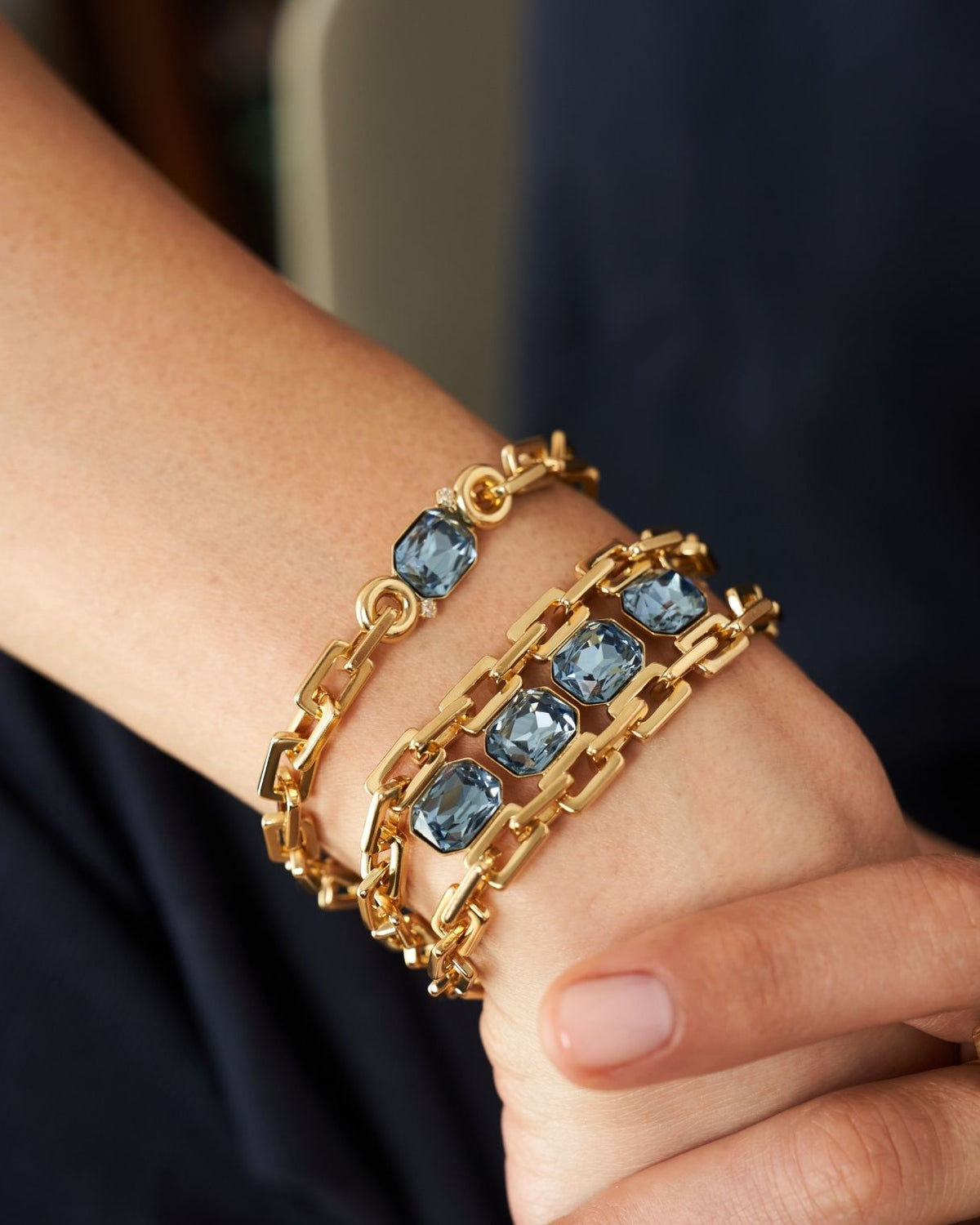 A close-up of a person's wrist wearing two stacked gold chain bracelets, each set with rectangular blue gemstones.