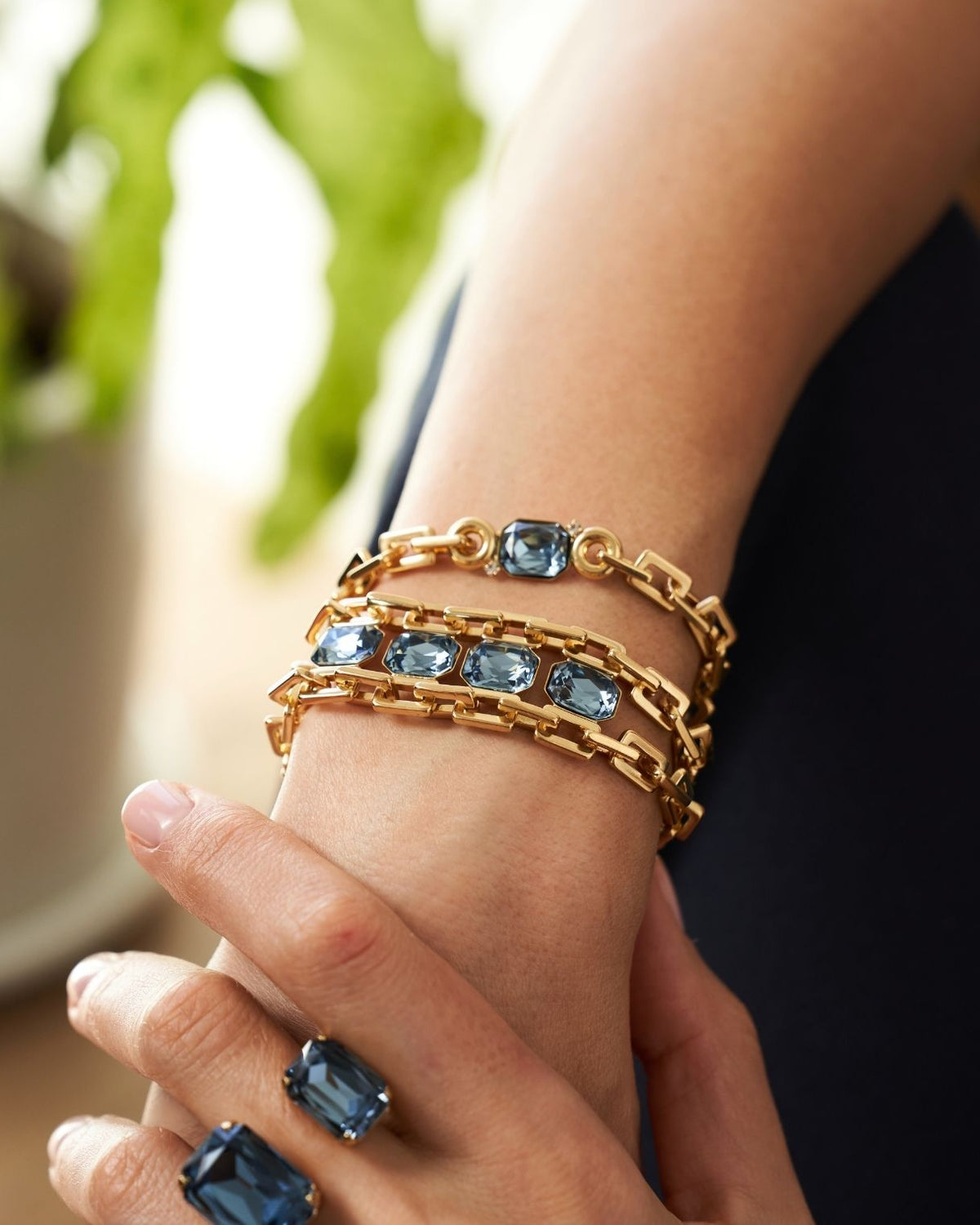 A close-up of a person's wrist and hand wearing multiple gold chain bracelets with rectangular blue gemstones, along with matching blue gemstone rings.